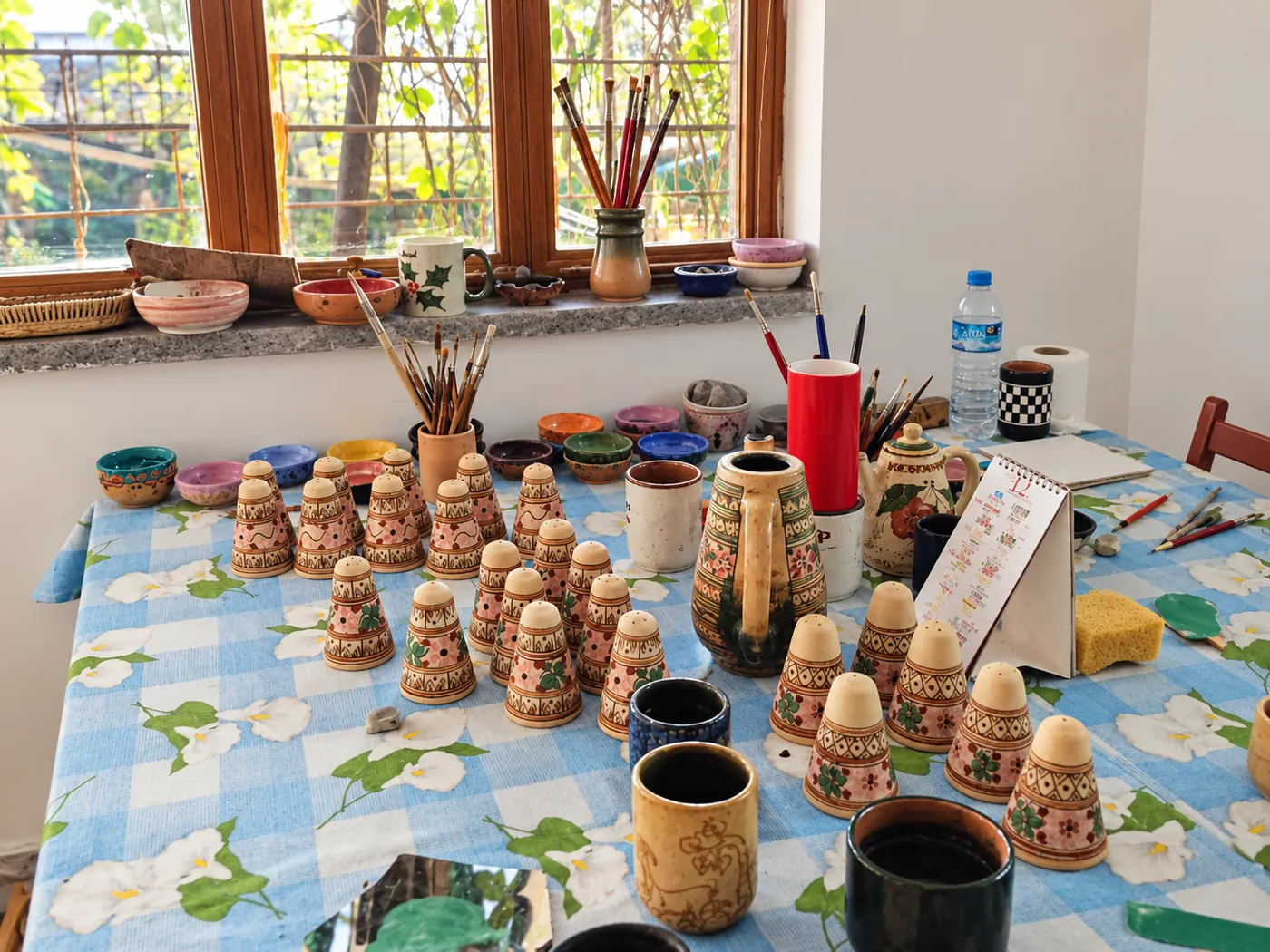 Studio painting table covered with freshly painted ceramic pieces, brushes and colourful glazes by the window